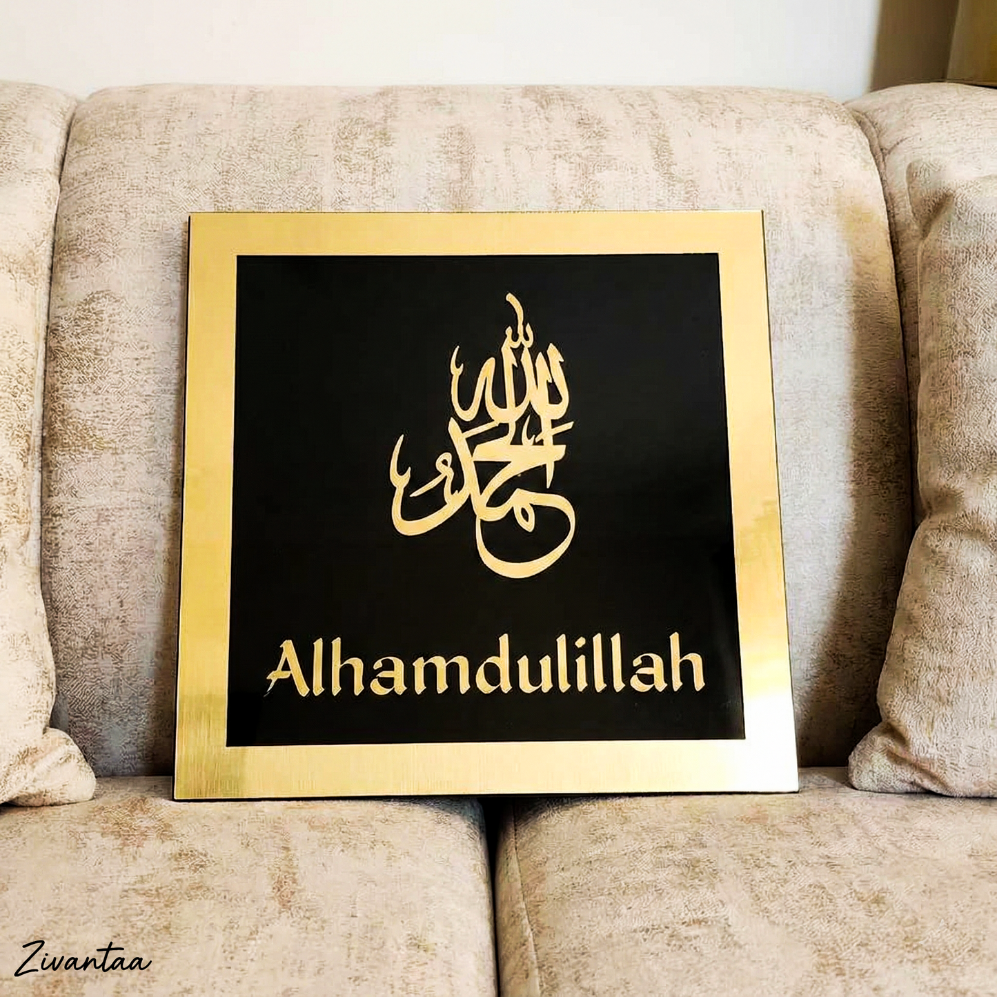 Decorative sign with 'Alhamdulillah' in gold on a black background, framed in gold, placed on a beige couch.