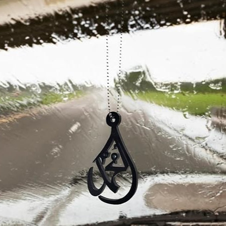 Decorative Arabic calligraphy pendant hanging from a car's rearview mirror on a rainy day.